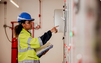 A construction worker in a blue hard hat and bright yellow coat pointing at an electrical box