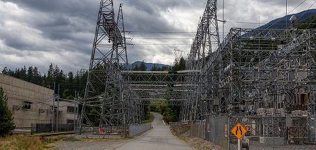 Electrical substation with metal towers and power lines, surrounded by forested hills and cloudy skies, featuring a narrow paved road running through the middle and a caution sign indicating aging infrastructure.